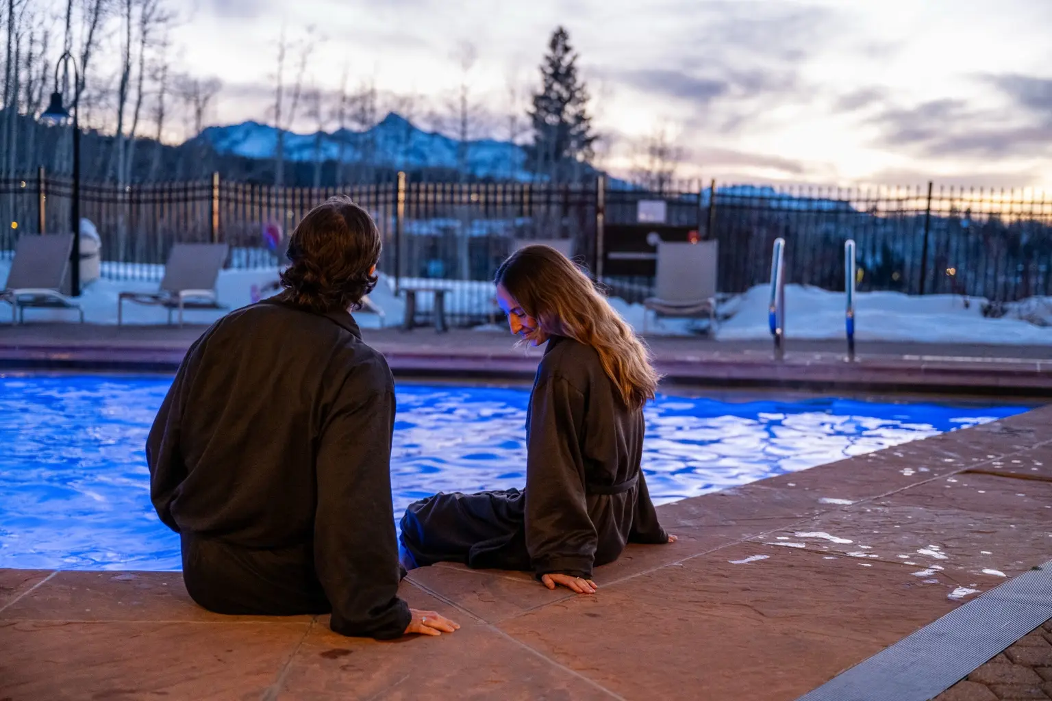 A couple in spa robes relax beside the outdoor pool at sunset in winter